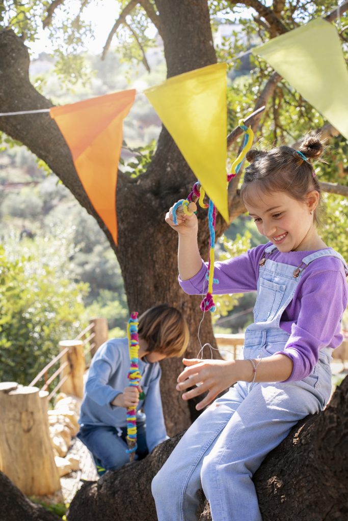 Idée d’atelier créatif facile à organiser pour animer une kermesse en plein air Fille jouant sous un arbre décoré de fanions colorés pendant une fête scolaire