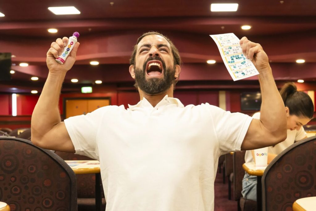 l'homme est dans une salle où se déroule un bingo loto. Homme criant victoire et brandit sa feuille de loto gagnante et un marqueur