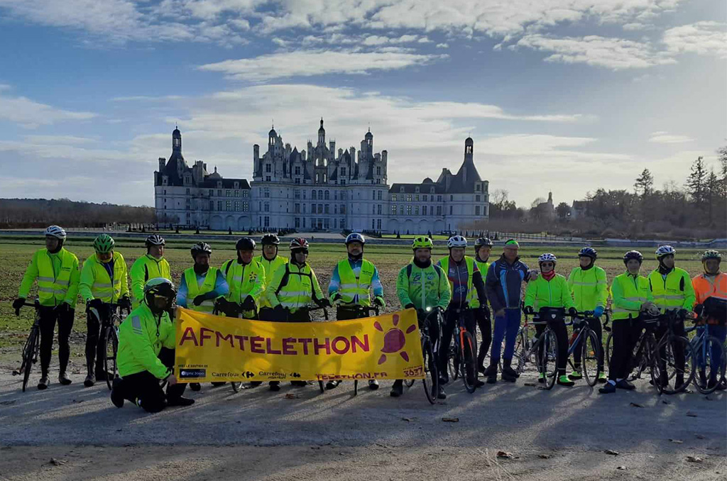 Cyclistes mobilisés pour le Téléthon lors d’une sortie devant un château Groupe de cyclistes du groupe EDF posant avec une banderole AFM Téléthon devant un grand château au loin